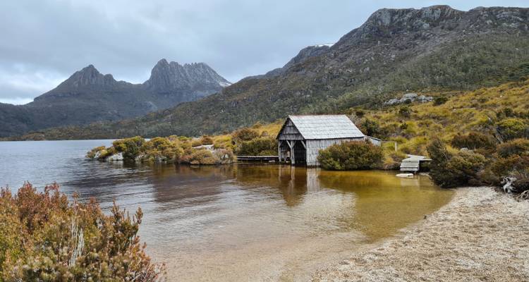 Serene alpine lake with weathered boatshed and jagged mountain skyline under overcast sky