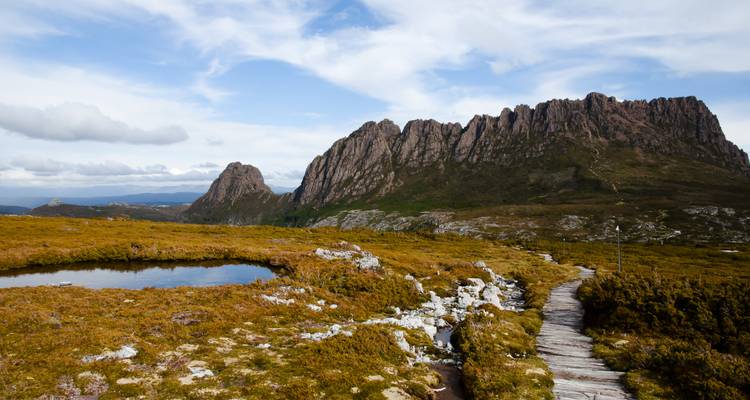 Alpine boardwalk winds past a small tarn toward a dramatic rocky ridgeline under patchy clouds