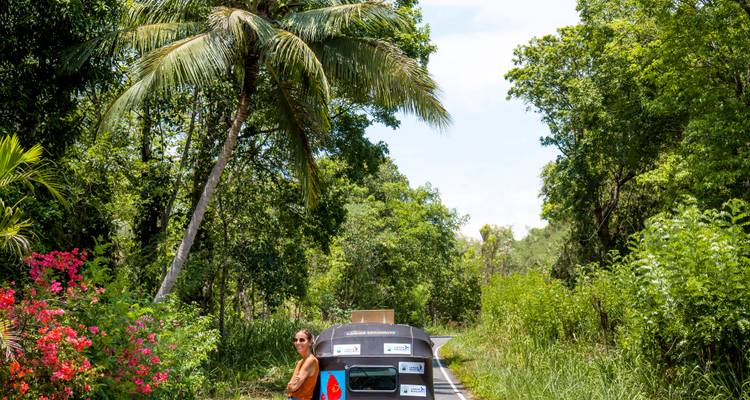 Reiziger die naast een zwarte tuk-tuk staat op een smal junglepad met een hoge palmboom erboven.