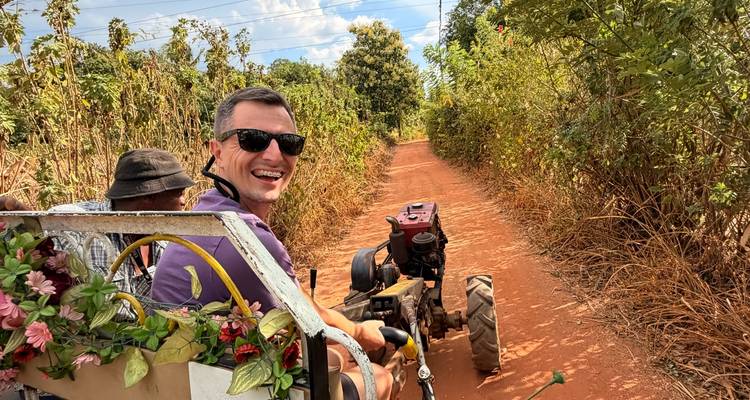 Reizigers glimlachen terwijl ze in een klein tractoraanhangertje over een smalle rode zandweg rijden, omzoomd door hoge droge struiken.