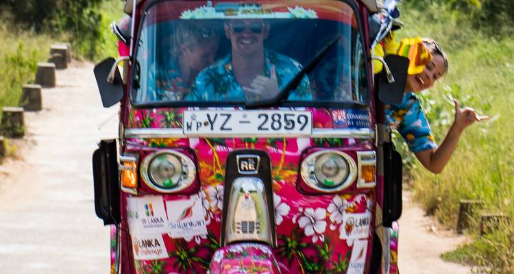 Een felroze tuk-tuk met bloemenpatroon rijdt op de camera af met lachende deelnemers, waarvan er één speels uit de zijkant posseert.