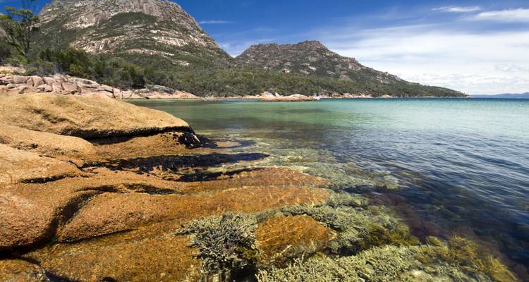 Crystal-clear water laps against orange granite rocks with forested mountains in the background.