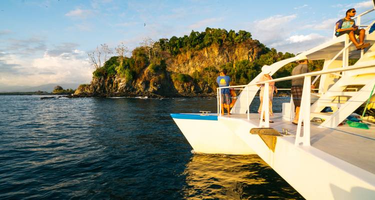 Les voyageurs se tenant sur le pont d'un bateau blanc s'approchant d'une petite île couverte de jungle à l'heure dorée.