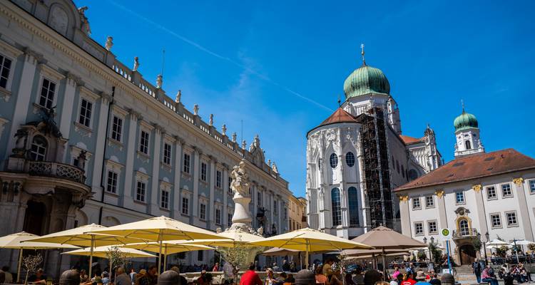 Plaza bulliciosa en Passau con edificios de color crema, sombrillas de cafetería y cúpula de la catedral.