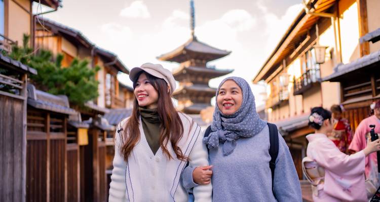 Twee jonge vrouwen lachen terwijl zij door een smalle traditionele straat slenteren met een vijfverdiepige pagode die in de achtergrond oprijst.