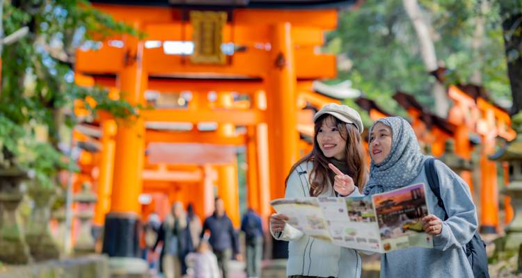 Twee vrouwelijke reizigers raadplegen een kaart en wijzen vooruit tussen levendige oranje torii-poorten, terwijl andere bezoekers wazig op de achtergrond zichtbaar zijn.