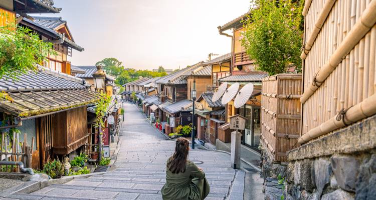 Een solo reiziger zit op stenen trappen en kijkt uit over een rustige historische steeg met traditionele houten machiya-huizen.