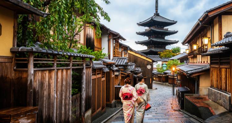 Twee vrouwen in bloemen kimono's lopen over een natte steeg naar een imposante pagode die omgeven is door houten stadhuizen.