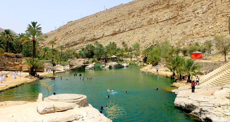 Natürliche Badebecken mit türkisblauem Wasser und entspannende Menschen unter felsigen Wüstenhängen in Wadi Bani Khalid.