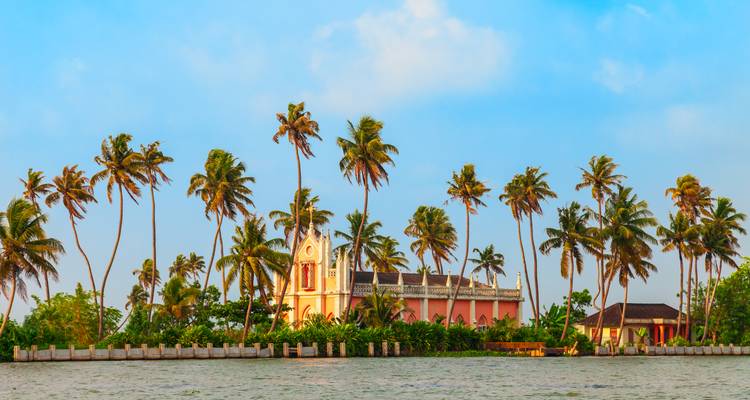 Line of swaying palm trees and a pastel church on a narrow strip of land in Kerala backwaters.