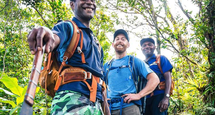 Three trekkers in tropical forest gear smile for the camera amid dense green jungle foliage.