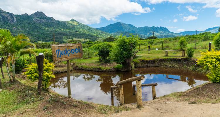 Small brown mud pool surrounded by greenery with rugged mountains forming a scenic backdrop and a wooden sign reading 'Mudpool'.