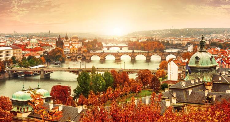 Sunrise view over Prague with the Vltava River lined by historic bridges and autumn-colored rooftops.