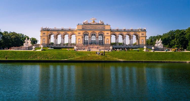Reflected view of the Gloriette across the still ornamental pond under clear blue sky.