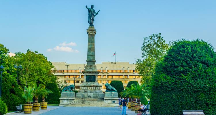 A tall freedom monument with bronze lions stands in a landscaped city square with visitors strolling nearby.