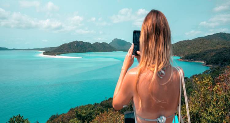 Back view of a woman photographing a sweeping turquoise bay and forested islands from a lookout