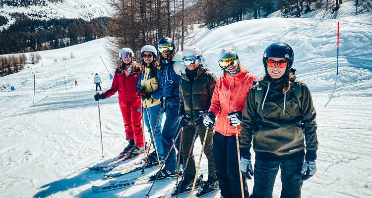 Eine Reihe von Freunden, die auf Skiern an einem sanften verschneiten Abhang unter strahlender Sonne posieren.