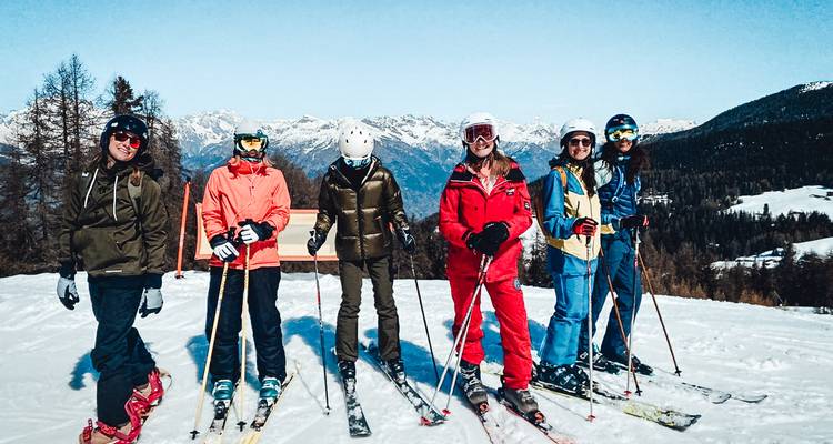 Gruppe von sechs Skifahrern, die auf einem Bergrücken mit breitem alpinem Panorama unter klarem Himmel lächeln.