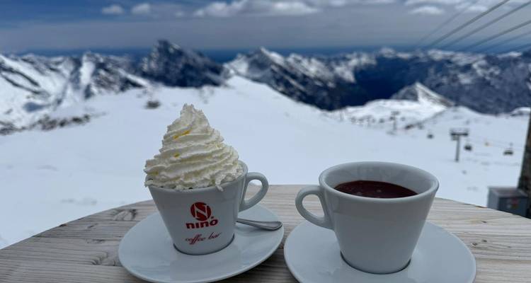 Zwei Tassen warme Getränke auf einem Holztisch mit Schlagsahne und Panoramablick auf verschneite Gipfel im Hintergrund.