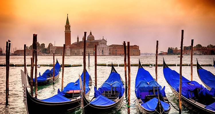 Row of blue-covered gondolas moored on the Venetian lagoon with San Giorgio Maggiore silhouetted at dawn.