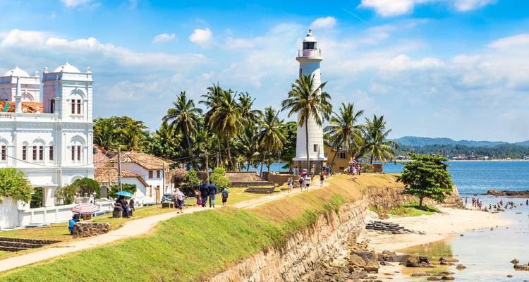 The white lighthouse on Galle Fort’s ramparts surrounded by palm trees and seaside visitors on a sunny day.
