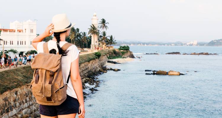 Female backpacker gazes over Galle’s seaside ramparts and lighthouse with waves and distant shore.