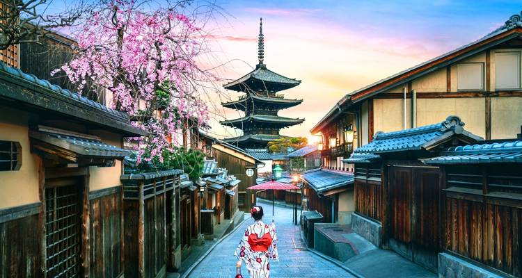 A kimono-clad visitor walks down a historic lane toward Kyoto’s Yasaka Pagoda as cherry blossoms frame the sunset sky.