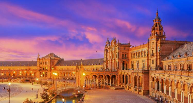 Der großartige Palastkomplex Plaza de España in Sevilla, beleuchtet durch warme Sonnenunterggangsfarben vor einem dramatischen purpur-orangefarbenen Himmel.