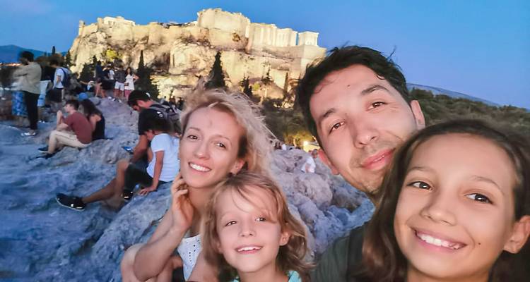 Abendliches Familienselfie auf einem felsigen Ausläufer mit der beleuchteten Akropolis im Hintergrund.