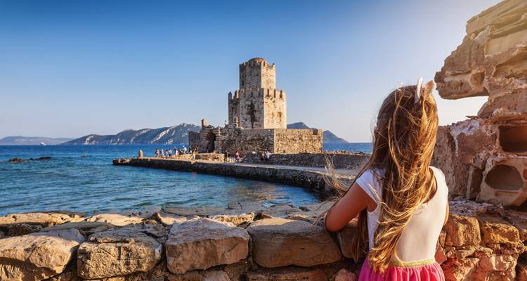 Une jeune fille contemple une forteresse en pierre en bord de mer depuis l'arrière d'un mur rustique.