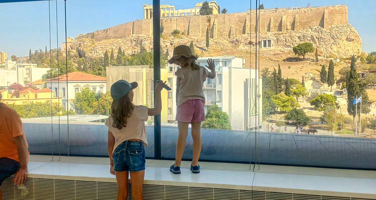 Two children press against a panoramic glass wall of the Acropolis Museum, gazing at the Acropolis hill outside.