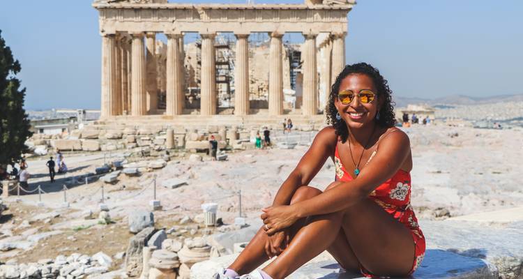 Glimlachende vrouw in zomeroutfit zittend op een marmeren plaat met het Parthenon achter haar.