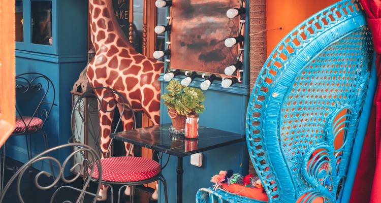Eclectische caféhoek met een levensgrote giraffe, een felblauw rieten stoeltje en retro spiegellampen.