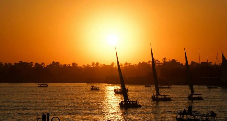 Traditional feluccas sail the Nile River silhouetted against a fiery orange sunset.