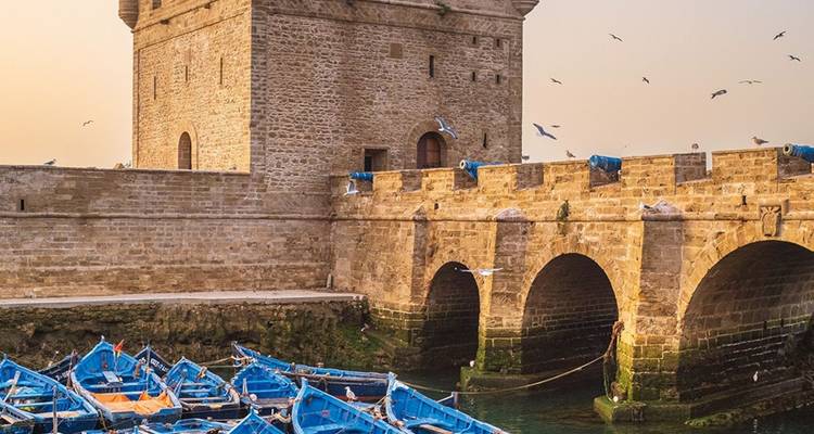 Stone sea fort and arched bridge with blue fishing boats in Essaouira harbor at dusk.