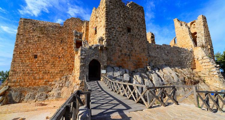 Fortaleza medieval de piedra bien conservada bajo un cielo azul brillante, a la que se accede por un puente de madera