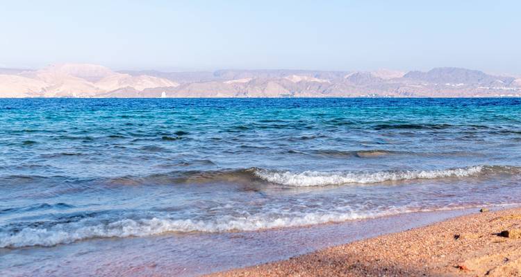Des vagues bleu clair qui s'écoulent doucement sur un rivage sablonneux avec des montagnes arides en arrière-plan