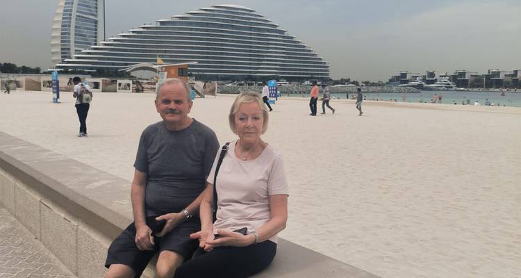 Un couple de personnes âgées est assis sur une promenade de plage avec le moderne hôtel Jumeirah Beach, de forme ondulante, s'élevant derrière eux.