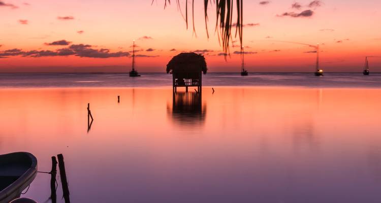 Lagon serein au coucher de soleil éclatant avec une petite paillotte sur pilotis et des voiliers ancrés en silhouette
