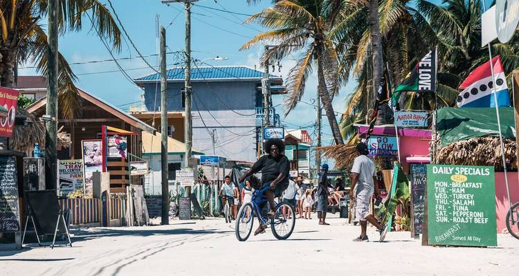 Un ciclista casual baja por la arenosa calle principal de Caye Caulker rodeado de señales coloridas y palmeras.