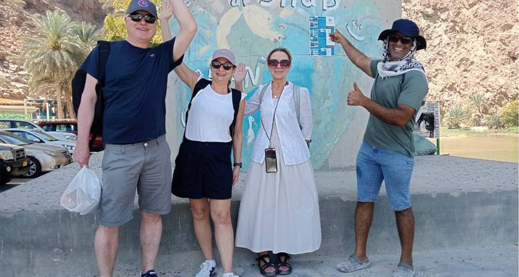 Des voyageurs heureux lèvent les mains et pointent du doigt vers une carte affichée sur un pilier accueillant les visiteurs à Wadi Shab.