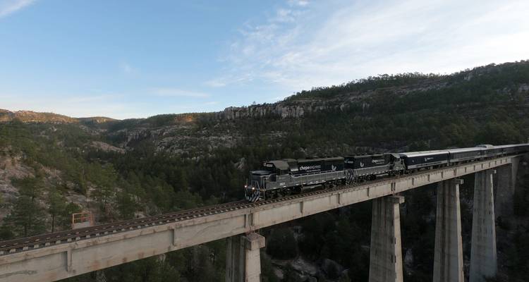 El tren Chepe Express cruza un puente elevado entre paredes de cañón cubiertas de bosque bajo un cielo pastel