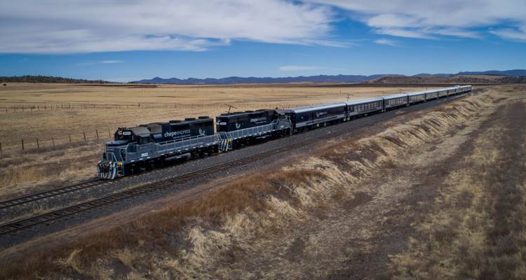 Vue aérienne d'un long train de voyageurs circulant à travers des plaines dorées ouvertes sous un ciel bleu.