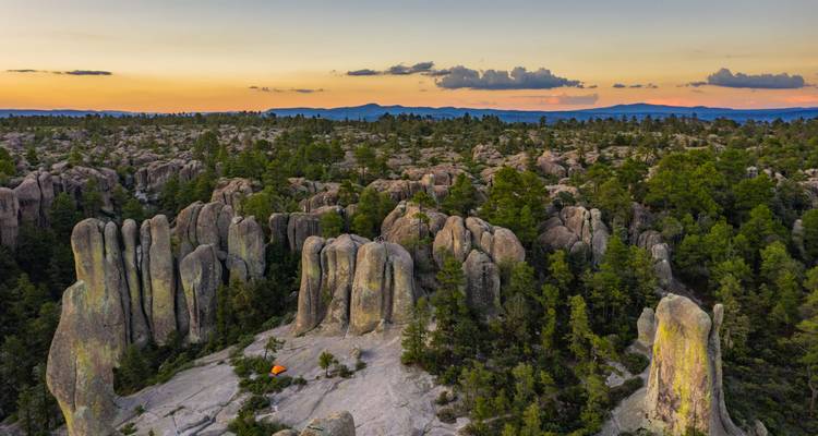Les pics rocheux et les pins brillent au coucher du soleil avec une tente orange solitaire dressée parmi les formations.