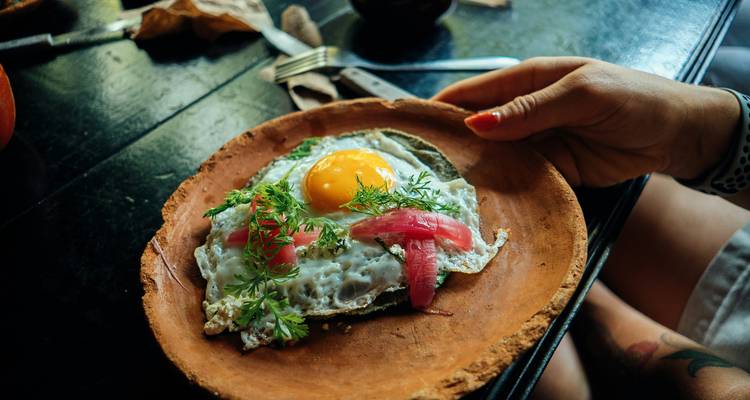 Assiette rustique en argile présentant un œuf au plat garni d'herbes et d'oignon mariné, une main la tenant sur une table en bois foncé.