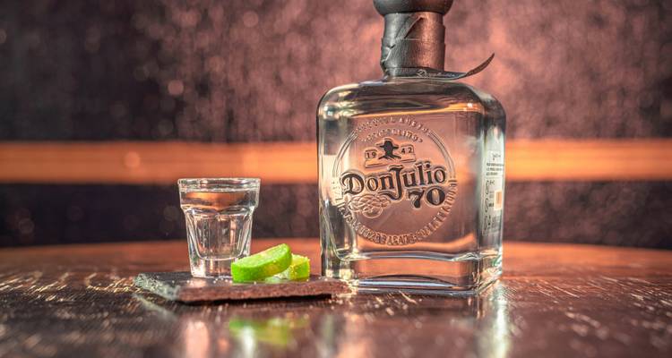 Bouteille de tequila Don Julio avec un verre à shot et des tranches de citron vert sur un bar en bois poli, légèrement éclairé.