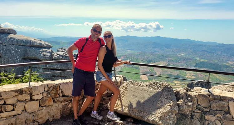 Couple stands at a rocky viewpoint overlooking rolling mountains and valleys under a clear sky.