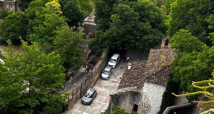 Vue aérienne d'une ruelle pavée avec des voitures garées à l'ombre de grands arbres dans une ancienne ville en pierre