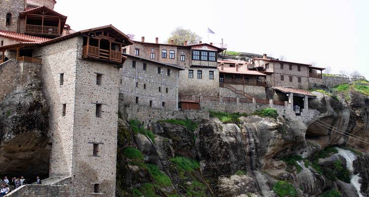 Panoramisch uitzicht op meerdere Meteora-kloosters die op ruwe rotsen liggen met bezoekers verzameld op het pad eronder.