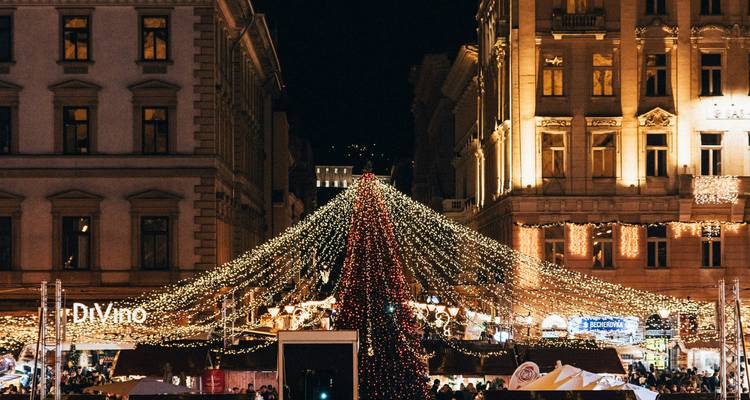Scène nocturne de bâtiments urbains encadrant un sapin de Noël illuminé s'élevant au-dessus d'un marché animé.